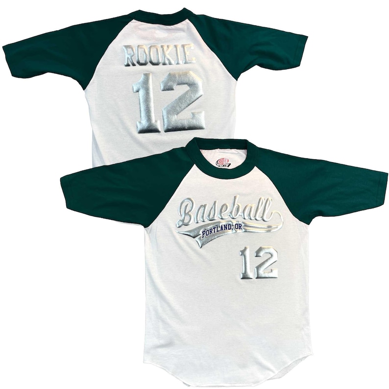 May include: White baseball jersey with dark green sleeves and collar. The jersey features the word "ROOKIE" and the number "12" in silver lettering on the back. The front of the jersey says "Baseball PORTLAND, OR" and the number "12".