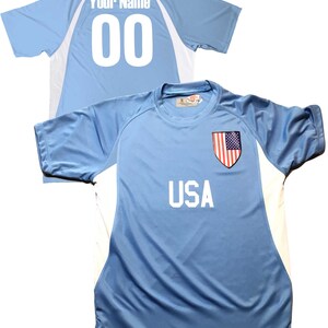 May include: A light blue soccer jersey with white accents. The front of the jersey has the word "USA" printed in white and a small American flag patch. The back of the jersey has the number "00" printed in white and the words "Your Name" printed in white above the number.