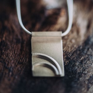 Op de afbeelding: Een zilveren ketting met een rechthoekige hanger met een gebogen ontwerp. De hanger ligt op een houten oppervlak.