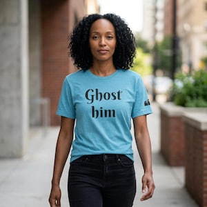 May include: A heathered teal short-sleeve t-shirt featuring the black text Ghost him printed on the chest. The shirt includes a small black graphic of two ghosts on the left sleeve and is paired with black denim jeans.