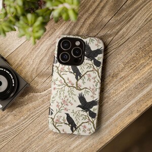 Puede incluir: Una funda de teléfono con un fondo crema con un patrón de cuervos negros posados en ramas florecientes con flores rosas. La funda está sobre una superficie de madera, con una cámara y vegetación en el fondo.