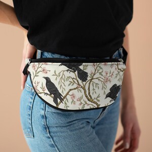 May include: A white fanny pack with a black zipper and strap. The pack features a repeating pattern of black crows perched on branches with pink blossoms and green leaves. The bag is worn around the waist.