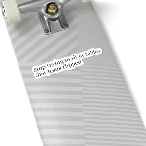 May include: A white sticker with the text "Stop trying to sit at tables that Jesus flipped" is placed on a skateboard deck. The deck has a gray and white striped pattern. The skateboard's metal trucks and wheels are visible.