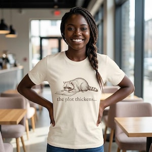 Puede incluir: Camiseta color crema con un mapache leyendo un libro y el texto "the plot thickens...". El mapache está en tonos grises y marrones. Una prenda casual.