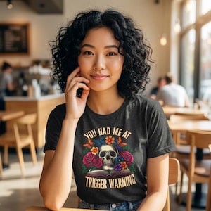 May include: Dark grey t-shirt featuring the text "YOU HAD ME AT TRIGGER WARNING" and a skull graphic with flowers and butterflies. The shirt is worn by a person.