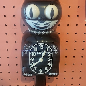 May include: Black Kit-Cat Klock in the shape of a cat with a white clock face. The clock has a rhinestone collar and accents. The clock face has black numbers and white hands. The clock is hanging on a pink pegboard.