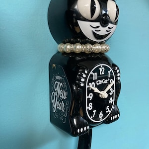 Può includere: Un orologio Kitt-Cat nero con una collana di perle. Il quadrante dell'orologio è bianco con numeri e lancette nere. Il lato dell'orologio recita "Happy New Year".