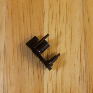 May include: Black plastic part with a small metal pin and a larger metal pin. The part has a rectangular shape with a small hole in the center.