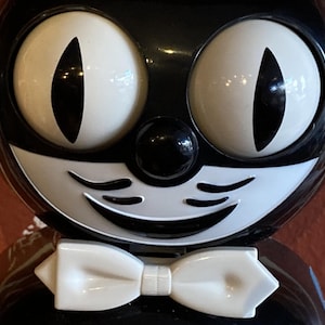 May include: A black and white cat figurine with large, round eyes and a white bow tie. The cat has a smiling mouth and a black nose.
