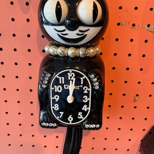 May include: A black Kit-Cat Klock wall clock featuring a white face with black numerals, large white eyes, and a wide smile. The cat wears a faux pearl necklace with silver-tone accents and has clear rhinestones embedded on its paws.