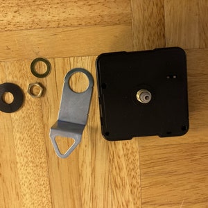 May include: Clock movement kit with black clock mechanism, silver metal mounting bracket, two black washers, and two gold-colored nuts.