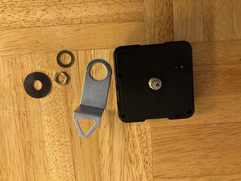 May include: Clock movement kit with black clock mechanism, silver metal mounting bracket, two black washers, and two gold-colored nuts.