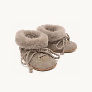May include: A pair of taupe suede ankle boots featuring a thick, fluffy shearling lining at the collar. The boots have a rounded toe, a flat tan sole, and thick, matching taupe rope laces that thread through metal eyelets across the front.