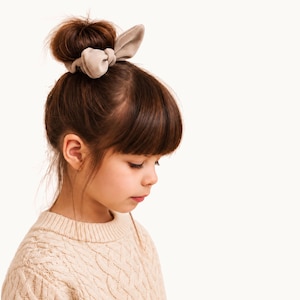 May include: A beige fabric hair scrunchie featuring two upright, rabbit-ear style fabric loops tied at the top. The scrunchie is worn around a high bun hairstyle, securing the dark brown hair in place against a plain, light-colored background.