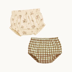 Peut inclure: Deux culottes bouffantes avec taille et ouvertures de jambes élastiques. L'une est de couleur crème avec un petit motif floral marron, et l'autre présente un motif à carreaux vichy marron et blanc. Les deux articles ont un bord volanté.