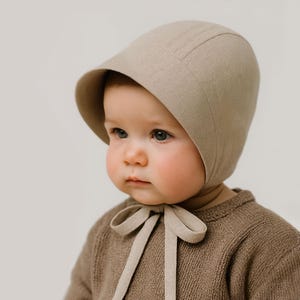 May include: A solid beige baby bonnet with a rounded crown and a wide brim that frames the face. The bonnet features matching fabric ties that fasten securely under the chin in a bow. The material has a smooth, matte texture and neutral tone.
