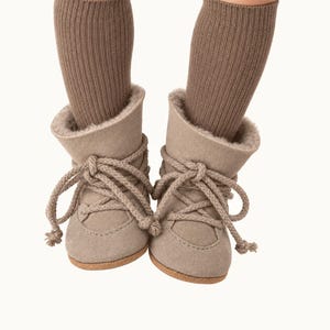 May include: A pair of beige suede ankle boots with thick, light brown rope laces and a soft, fuzzy interior lining. The boots are worn with ribbed, knee-high brown socks that extend upward from the top of each boot, creating a layered appearance.