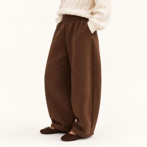 May include: Dark brown sweatpants featuring a high-rise elastic waistband and a loose, wide-leg fit. The pants are paired with a cream-colored cable knit sweater and dark brown ballet flats, creating a simple and cohesive outfit ensemble.