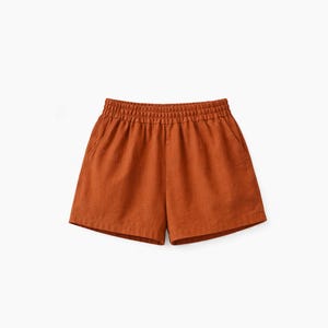 May include: Rust-colored shorts featuring an elasticized waistband with gathered fabric detail. These shorts have a relaxed fit, a straight hemline, and two side pockets, providing a simple and functional design for casual wear.
