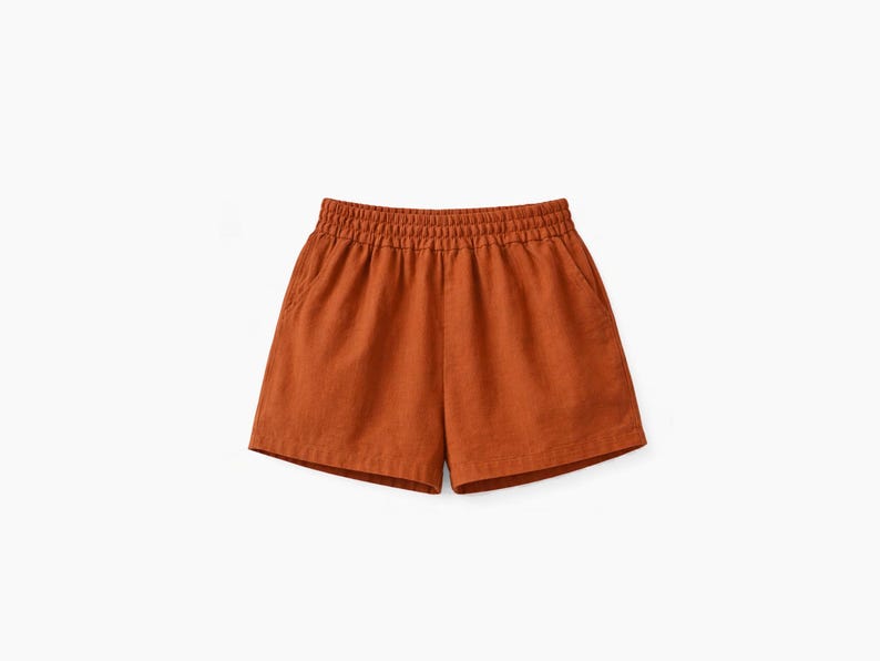 May include: Rust-colored shorts featuring an elasticized waistband with gathered fabric detail. These shorts have a relaxed fit, a straight hemline, and two side pockets, providing a simple and functional design for casual wear.