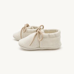 May include: A pair of off-white, soft-textured baby booties featuring a rounded toe design and matching beige fabric laces tied in a bow at the front. The booties have a simple, slip-on construction with a flexible sole and a clean, neutral appearance.