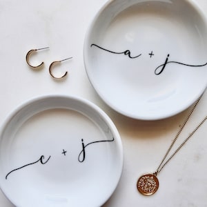 May include: Two white ceramic ring dishes with black calligraphy. The dishes are labeled "a + j" and "c + j". A gold necklace with a round pendant is on a white surface.