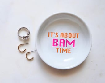 May include: A white ceramic trinket dish with the text "IT'S ABOUT BAM TIME" in orange and pink. A silver ring and two gold hoop earrings are next to the dish. The items are on a white surface.