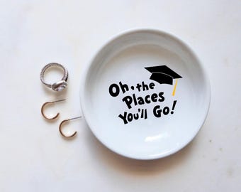 May include: White ceramic ring dish with a black graduation cap and the text "Oh, the Places You'll Go!" printed on it.