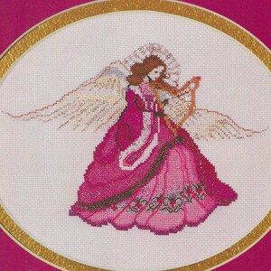 May include: A cross-stitch pattern featuring a Baroque Angel playing a harp. The angel is wearing a pink dress with gold trim and has a halo. The pattern is framed in a gold circle on a pink background. The text "Baroque Angel" and "Mar Bek" are printed below the image.