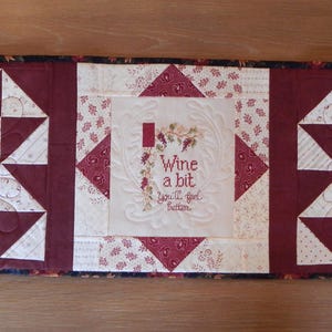 May include: A red, white, and black quilted table runner with a central panel featuring the text "Wine a bit" in white embroidery. The runner is decorated with a repeating pattern of white and red triangles.