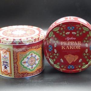 May include: Two decorative red and gold cookie tins. One is round with a floral design and the text "Peppar Kakor". The other is cylindrical with a floral and geometric pattern. Both tins are likely for storing cookies.