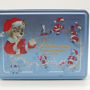 May include: A blue metal tin with a Christmas theme. It features a cartoon bear dressed as Santa Claus holding a gift box with fireworks coming out of it. The text on the tin reads "Delivering Christmas magic since 1926".