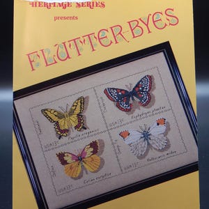 May include: A craft book cover with the title "FLUTTER-BYES" in red letters. The cover features a framed cross-stitch design of four butterfly stamps, each with a different species name and "USA 13C" text. The background is yellow.