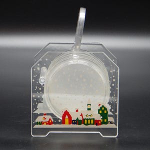 May include: A clear acrylic coaster set with a winter village scene. The holder has a handle and a decorative border with colorful houses and falling snow. The set includes several round coasters.