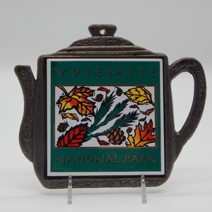May include: A brown metal teapot-shaped trivet with a square tile insert. The tile features a green background with the text "Yosemite National Park" and a colorful illustration of autumn leaves and pine branches.