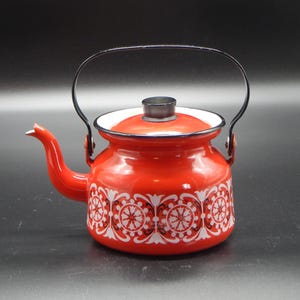 May include: A bright red enamel tea kettle featuring a white and black floral pattern around the base. The kettle has a black metal handle, a matching red lid with a black knob, and a curved spout. The interior rim of the lid is white.