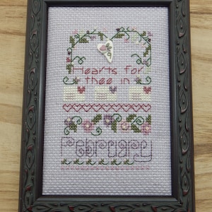 May include: A framed cross-stitch sampler with a purple background and pink and green floral accents. The sampler reads "Hearts for thee in February".