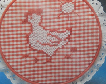 May include: A red and white gingham fabric hoop with a white embroidered chicken and sun design. The hoop has a white lace trim and a red and white gingham ribbon tied around it.
