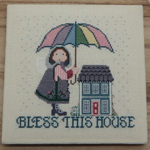 May include: A cross-stitch embroidery featuring a girl holding a colorful umbrella over a small house. The text "Bless This House" is stitched below the image.