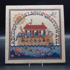 May include: A cross-stitched square featuring Noah's Ark with animals, a sun, and the alphabet. The ark is red-roofed, with pairs of animals. The border is a checkered pattern in blue and beige. The background is black.