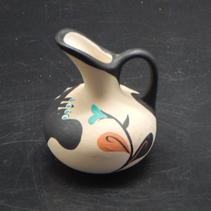 May include: A small, cream-colored ceramic pitcher with a black handle and spout. The pitcher is decorated with black, turquoise, and orange floral designs. The pitcher is approximately 5 inches tall.