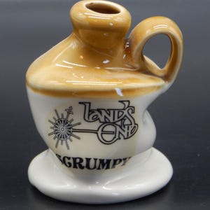 May include: A small ceramic jug with a brown glaze and a white base. The jug has a handle and a spout. It features a black and white logo that says "Land's End" and "Grumpy".