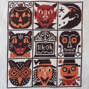 May include: A cross-stitch sampler featuring Halloween-themed designs. The sampler includes a jack-o'-lantern, devil, crescent moon, black cat, tombstone with "Uh Oh" text, skull, owl, witch, and scarecrow. The color palette is primarily orange, black, and brown.