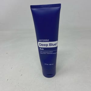 Puede incluir: Un tubo de crema azul oscuro de dōTERRA Deep Blue Rub. La etiqueta blanca muestra el texto dōTERRA, Deep Blue, Rub, with proprietary CPTG Deep Blue essential oil blend y 4 fl oz / 120 mL. El tubo tiene una tapa azul oscuro a juego en la base.
