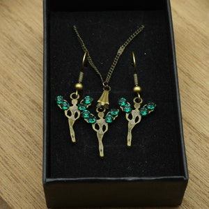 Puede incluir: Un conjunto de joyas que incluye un collar y pendientes. El collar y los pendientes están hechos de bronce y tienen un diseño de cristal verde. Las joyas se muestran en una caja negra.