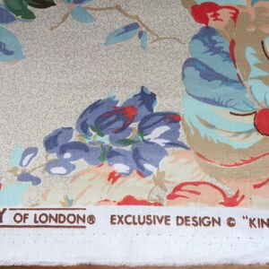 May include: Close-up of floral fabric featuring large, colourful flowers in red, blue, and peach hues against a beige backdrop. The fabric is printed with "LIBERTY OF LONDON" and "EXCLUSIVE DESIGN &copy; "KINTBURY"". Ideal for dressmaking or crafting.