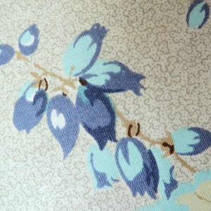 May include: Close-up of a fabric with a floral pattern. The design features blue and teal-coloured stylized flowers and leaves on a beige background. The fabric has a textured appearance, suggesting a woven material.
