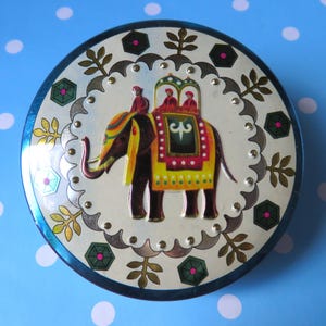 May include: A round, decorative tin with a blue rim and a white center featuring an ornate elephant design. The elephant is adorned with colorful patterns and carries three figures. The tin is surrounded by a decorative border with floral and geometric elements.
