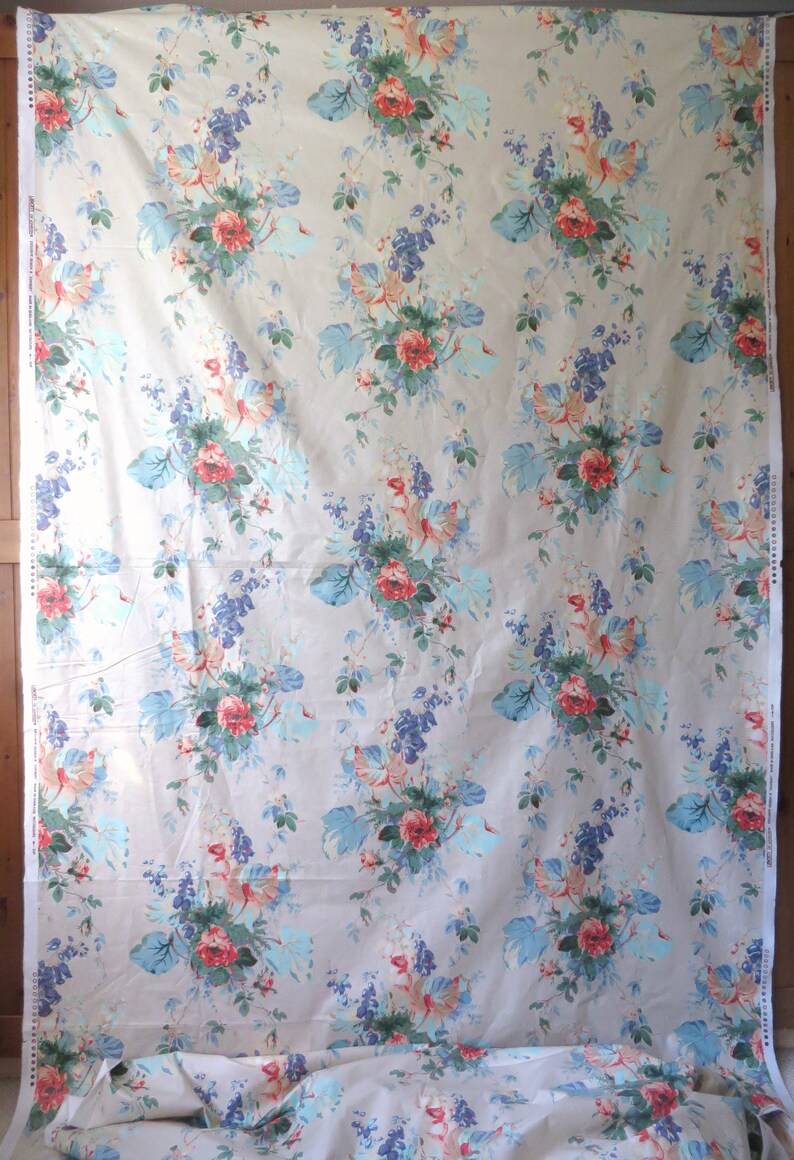 May include: A large piece of fabric with a repeating floral pattern. The design features clusters of red roses, blue and purple flowers, and large, light blue leaves on a beige background. The fabric is draped.