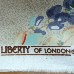 May include: Close-up of Liberty of London fabric. The fabric displays a floral pattern with blue, teal, and peach flowers on a beige backdrop. The brand name is printed in brown letters.